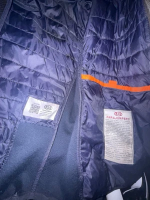 Mörkblå pufferjacka från Parajumpers - Snygg mörkblå pufferjacka från Parajumpers med klassisk quiltad design och orange detalj på insidan. Jackan har lätt glansigt yttertyg och är perfekt för kalla vinterdagar. Märket syns tydligt på inneretiketten och jackan har en modern, sportig look.