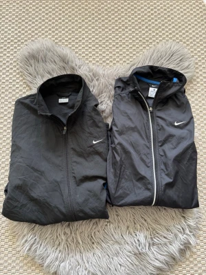 2-Pack Svarta Nike vindjackor med detaljer - Två svarta vindjackor från Nike med dragkedja och vita detaljer. Ena jackan har blå inslag på sidorna och huva, den andra har vita ränder längs ärmarna. Båda har Nike-logga på bröstet och praktiska fickor. Perfekta för sport eller chill dagar.