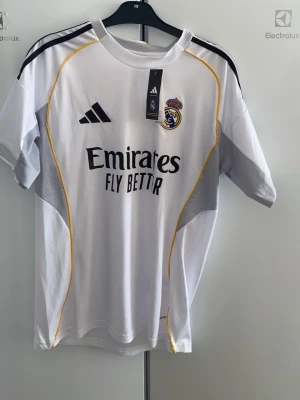 Real Madrid hemmaställ säsong 25/26 - Oanvänd och snygg vit Real Madrid matchtröja från Adidas med svarta och gula detaljer. Tröjan har klubbmärke och Adidas-logga på bröstet samt 'Emirates Fly Better' tryckt framtill. Tillverkad i lätt och ventilerande material, perfekt för fotbollsträning, match eller en avslappnad vardagsstil. 