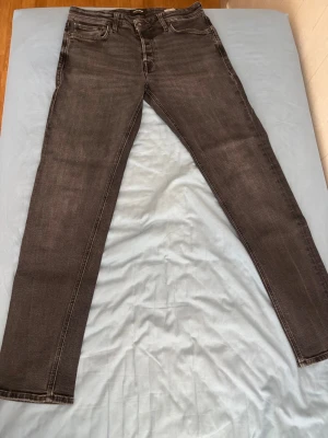 Svarta tapered jeans Jack & Jones 32/34 - Snygga svarta jeans från Jack & Jones i modellen Tapered/Mike. Jeansen har fem fickor, normal midja och avsmalnande ben. Klassisk knappgylf och diskreta slitningar för en cool look. Perfekta för dig som gillar en avslappnad men stilren stil.