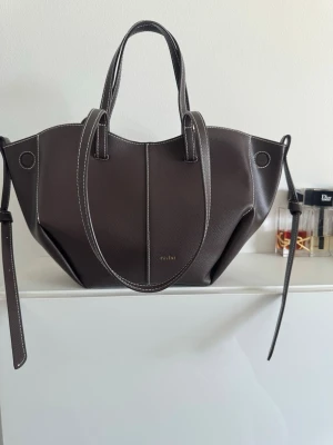 Polène brun handväska i läder - Snygg brun handväska från Polène i strukturerat läder med vita kontrastsömmar. Väskan har en unik, skulptural form med dubbla handtag och långa sidoremmar. Perfekt för dig som gillar stilrena och moderna accessoarer. Cyme mini bag färgen enoby