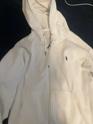 Vit hoodie från Polo Ralph Lauren - Snygg vit hoodie från Polo Ralph Lauren med dragkedja framtill och klassisk broderad logga på bröstet. Hoodien har huva med dragsko och två fickor framtill. Perfekt för en clean och stilren look. Priset kan diskuteras.