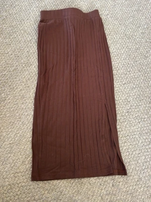 Brun ribbad långkjol - Säljer en brun långkjol med ribbad struktur och slits på sidan. Kjolen har en enkel och stilren design som passar perfekt till olika outfits. Materialet känns mjukt och följsamt, vilket gör den bekväm att bära hela dagen.