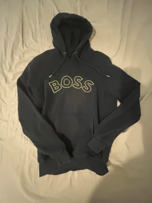 Mörkblå hoodie från BOSS - Snygg mörkblå hoodie från BOSS med stor vit logotyp på bröstet. Hoodien har en klassisk huva med dragsko och en stor magficka. Perfekt för dig som gillar streetwear och vill ha något bekvämt och stilrent. Materialet är mjukt och känns riktigt skönt.