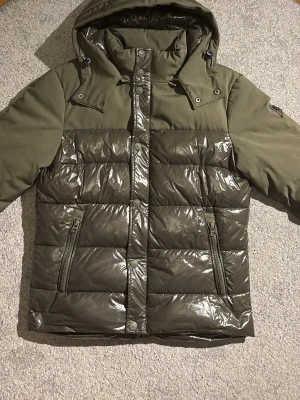 Olivgrön pufferjacka från Invicta - Snygg olivgrön pufferjacka från Invicta med glansig vadderad front och matta ärmar. Jackan har AVTAGBAR huva, dragkedjefickor och tryckknappar framtill. Perfekt för kalla vinterdagar. Jackan har ett litet stygn i luvan.