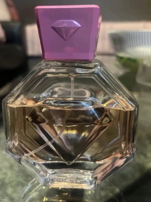 I använt skick. Parfymflaska med diamantmönster - Snygg parfymflaska i transparent glas med diamantformad design och ljuslila lock. Flaskan har en geometrisk form och innehållet är ljusrosa. Perfekt för dig som gillar unika och trendiga doftflaskor.