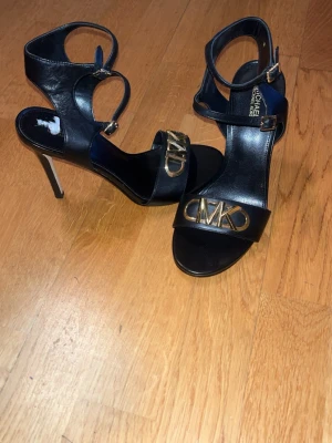 Svarta klackar från Michael Kors - Snygga svarta klackar i läder från Michael Kors med höga stilettklackar och guldfärgad MK-logga framtill. Skorna har dubbla remmar med spännen runt vristen för extra support och en elegant, öppen tå. Storlek 37 passar 38