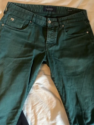 Mörkgröna jeans från Scotch & Soda - Snygga mörkgröna jeans från Scotch & Soda med klassisk femficksdesign och läderpatch bak i midjan. Jeansen har raka ben och är tillverkade i ett mjukt bomullsmaterial med lite stretch för extra komfort. Perfekta för dig som vill sticka ut med färg. W30/L34