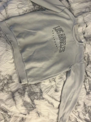 Ljusgrå sweatshirt Los Angeles - Ljusgrå sweatshirt med trycket 'Los Angeles California' på bröstet. Tröjan har rund halsringning, ribbade muddar vid ärmslut och nederkant samt en avslappnad passform. Perfekt för en casual streetwear-look.