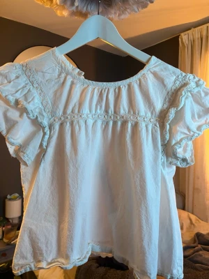 Vit blus med volang från Vero Moda - Supersöt vit blus från Vero Moda med korta volangärmar och spetsdetaljer. Blusen har en lös passform och rund halsringning, perfekt för en fräsch och luftig look. Materialet är mjukt och lätt, vilket gör den bekväm att bära hela dagen.