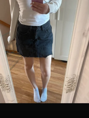 Svart jeanskjol med rå kant - Säljer en svart jeanskjol med rå, fransad kant nertill och klassiska fickor fram och bak. Kjolen har en rak passform och är i ett mjukt denimtyg. Perfekt för dig som gillar en enkel men edgy stil.