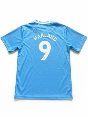 Manchester City Hemmatröja 23/24 – HAALAND #9 (Junior L) - Säljer nu den snygga hemmatröjan från Manchester Citys säsong 23/24. Tröjan pryds av världsstjärnan Erling Haaland med nummer 9 på ryggen. Designen är inspirerad av Etihad Stadiums 20-årsjubileum med de karaktäristiska ränderna i tyget. • Spelare: Erling Haaland • Storlek: Junior L (motsvarar ca 147–158 cm / 12–13 år). • Viktigt: Observera att detta är en barnstorlek, inte vuxenstorlek. • Skick: Mycket fint skick! Inga revor eller tydliga fläckar, och trycket sitter bra.