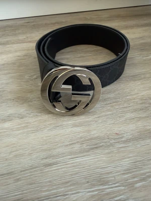 Svart Gucci bälte !!ÄKTA!! - Snyggt svart bälte från Gucci med en stor, rund metallspänne med GG-logga i silver. Bältet har diskret mönster längs remmen och känns riktigt lyxigt. Perfekt accessoar för att lyfta vilken outfit som helst. Den är 100cm, säljer pågrund av att den blivit för liten, går att byta mot 110cm/ 105