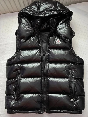 Moncler Bormes  - Snygg svart dunväst från Moncler med glansig finish, huva och dragkedja framtill. Västen har två praktiska fickor med dragkedja och klassisk Moncler-logga på bröstet. Perfekt för lager-på-lager och streetstyle.