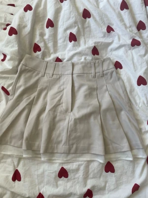 Beige plisserad kortkjol - Snygg beige kortkjol med plisserad design och breda hällor i midjan. Kjolen har en klassisk och stilren look som passar perfekt till många olika outfits. Materialet känns mjukt och bekvämt mot huden.