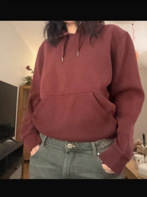 Vinröd hoodie med magficka - Säljer en vinröd hoodie med huva och dragsnören. Hoodien har en stor magficka framtill och ribbade muddar vid ärmslut och nederkant. Perfekt för en avslappnad och chill stil.