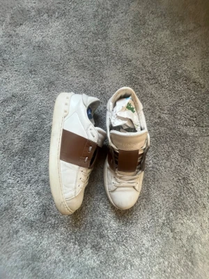 Vita Valentino sneakers med brun detalj - ÄKTA Valentino skor  med bred brun läderdetalj över vristen. Klassisk låg modell med rund tå och vita snören. Skorna har en stilren design och är tillverkade i läder för en exklusiv känsla. DUST BAG MEDFÖLJER!