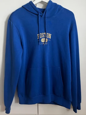 Pier one hoodie  - Blå hoodie med huva och dragsko, samt magficka framtill. På bröstet finns ett gult och vitt tryck med texten 'BOSTON USA 1992'. Perfekt för en avslappnad och sportig stil.
