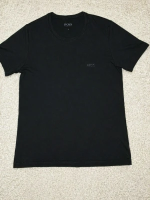 Svart t-shirt från BOSS - En stilren svart t-shirt från BOSS med diskret logga på bröstet. Klassisk rund halsringning och korta ärmar. Perfekt basplagg i garderoben och lätt att matcha med allt. Tillverkad i mjuk bomull för skön komfort.