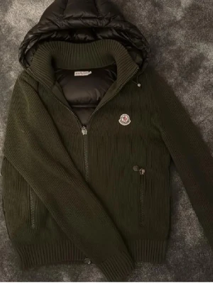 Olivgrön Moncler stickad hoodie - 3 är ungefär M är 186 och den passar klockrent. Snygg olivgrön stickad hoodie från Moncler med quiltad svart huva och dragkedja. Tröjan har ribbade muddar, två fickor med dragkedja och Moncler-logga på bröstet. Perfekt för dig som vill ha en cool och exklusiv look. Riktigt fett köpt för 9000. Lite färg på sidan men inget som märks när man använder cardiganen. Går och få ner i pris vid snabbaffär.