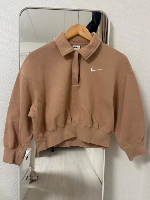 Beige cropped sweatshirt från Nike - Säljer en beige cropped sweatshirt från Nike med klassisk vit swoosh-logga på bröstet. Tröjan har krage och halv dragkedja framtill, samt breda muddar vid ärmslut och midja. Perfekt för en avslappnad och sportig stil.
