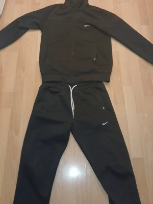 Svart mjukisdress  från Nike - Svarta mjukisbyxor från Nike med vit broderad logga på benet och liten Nike-tag vid fickan. Byxorna har dragsko i midjan och är tillverkade i mjukt material för maximal komfort. Perfekta för en avslappnad stil.