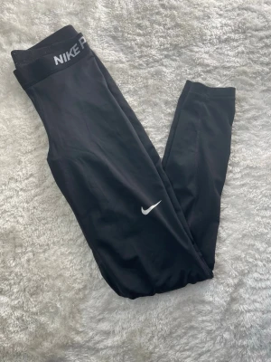 Svarta Nike Pro träningstights - Svarta träningstights från Nike Pro med elastisk midja och vit Nike-logga på benet. Tight passform och stretchigt material som passar perfekt för gym, löpning eller annan träning. Snygg och sportig design med diskret branding.