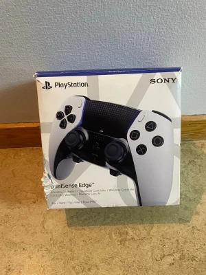 PlayStation DualSense Edge handkontroll - DualSense Edge är en trådlös handkontroll till PlayStation 5 från Sony. Den har en vit och svart design med ergonomiska grepp, justerbara knappar och avancerade funktioner för gaming. Materialet är plast med texturerad yta för extra komfort.