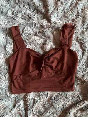 Brun croppad linnetopp med twist - Snygg brun croppad linnetopp med breda axelband och twistad detalj framtill. Toppen har en enkel, stilren design och är gjord i mjuk bomull som känns skön mot huden. Perfekt att matcha med höga jeans eller kjol för en trendig look.