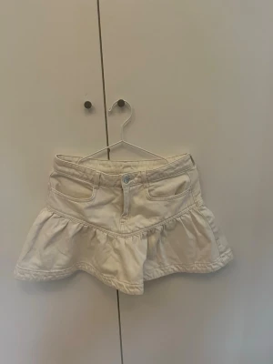 Vit volangkjol med fickor - Söt vit kortkjol med volangkant och två framfickor. Kjolen har en klassisk jeanslook med knapp och dragkedja framtill. Perfekt för dig som gillar en avslappnad men trendig stil.