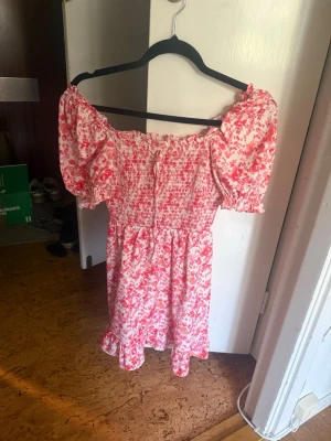 Blommig offshoulder klänning rosa/vit - Söt kort offshoulder klänning med puffärmar i vitt och rosa blommönster. Smockad överdel och volangkant nedtill ger en romantisk känsla. Perfekt för varma dagar och sommarens alla tillfällen.