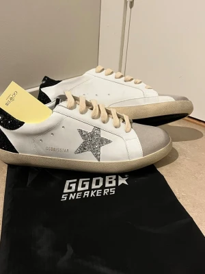 GGDB vita sneakers med glitterstjärna - Snygga vita sneakers från Golden Goose med en glittrig silverstjärna på sidan och svart hälparti med glitter. Skorna har beige snörning och tåparti i ljus mocka. Klassisk låg modell med vit sula och GGDB-logga på sidan.