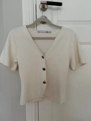Beige v-ringad topp från lager 157 - Snygg beige topp från lager 157 med v-ringning och korta ärmar. Toppen har tre dekorativa knappar framtill och är i ett mjukt, stretchigt material som känns skönt mot huden. Perfekt till jeans eller kjol för en clean look.