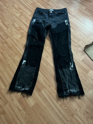 Svarta jeans med slitningar - Unika svarta bootcut jeans med råa kanter och slitningar framtill. Jeansen har vita färgstänk och detaljerade sömmar som ger en edgy vibe. Perfekta för dig som vill sticka ut med din stil. Materialet är klassisk denim. Aldrig använda 