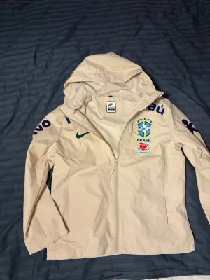 Nike Brasil vindjacka med huva - Stilren beige vindjacka från Nike med Brasiliens fotbollslandslagslogga på bröstet, grönt Nike-märke och flera tryck i blått och lila. Jackan har hel dragkedja, huva och sidofickor. Perfekt för dig som gillar sportig stil och vill visa ditt stöd för Brasilien. Helt ny står M men är liten för den stolek skulle passa som en liten S skulle säga. Pris kan diskuteras