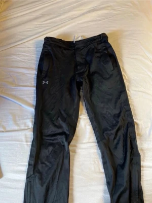 Svarta träningsbyxor från Under Armour - Snygga svarta träningsbyxor från Under Armour med diskret logga på vänster lår. Byxorna har raka ben, resår i midjan och sidofickor. Perfekta för gymmet eller löprundan. Materialet är lätt och andas bra.