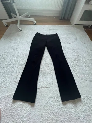Svarta kostymbyxor med utsvängda ben - Snygga svarta kostymbyxor med utsvängda ben och låg midja. Byxorna har klassisk passform och stängs med dragkedja och knapp framtill. Perfekta för en stilren look och passar till många olika outfits.