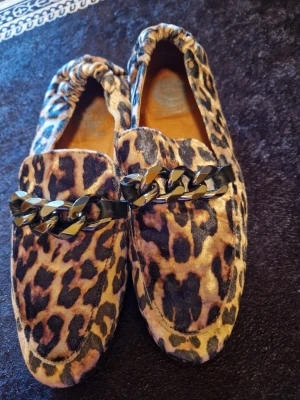 Leopardmönstrade loafers med kedja - Säljer ett par loafers från Forever 21 med leopardmönster i brunt och svart. Skorna har en chunky kedjedetalj i svart metall över foten och är tillverkade i ett mjukt tygmaterial. Perfekta för dig som vill sticka ut med din stil.