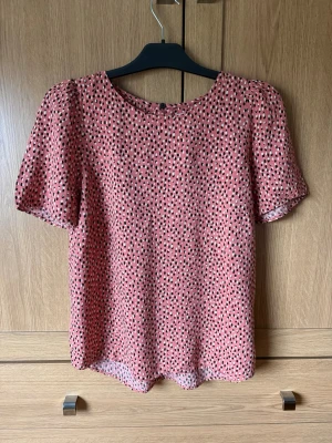 Kortärmad blus - Söt blus i korallrosa med svart och vitt prickigt mönster. Blusen har rund halsringning, lätt puffade axlar och korta ärmar. Tillverkad i ett tunt, luftigt tyg som ger en avslappnad och trendig look. 