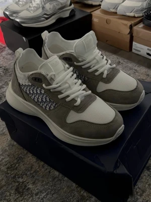 Dior B27 sneakers i grå och vitt - Snygga Dior B27 sneakers i grå mocka och vitt mesh med ikoniskt Dior Oblique-mönster på sidorna. Skorna har vita snören, chunky sula och en modern siluett som passar perfekt till streetwear. Kommer med originalförpackning och Dior-detaljer.