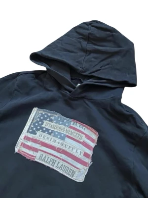 Svart hoodie Ralph Lauren denim & supply - Svart hoodie från Ralph Lauren med stor huva och känguruficka. På bröstet finns ett tryck med amerikansk flagga och texten 'Denim & Supply Ralph Lauren'. Tillverkad i mjukt bomullsmaterial och har långa ärmar. Perfekt för en avslappnad streetwear-look.