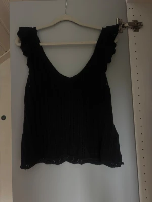 Svart stickad ärmlös topp - Snygg svart stickad topp med v-ringning och volangdetaljer vid axlarna. Toppen är ärmlös och har ett luftigt, stickat/virkat  material som passar perfekt till varma dagar. Enkel att styla med både jeans och kjol. Använd kanske 2 gånger