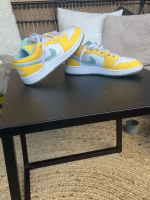 Nike Air Jordan 1 Low gul och mint - Säljer ett par Nike Air Jordan 1 Low sneakers i vitt, gult och mintgrönt. Skorna har klassisk låg siluett, vit bas med gula overlays och mintgrönt swoosh samt innersula. Tillverkade i läder med vita skosnören och detaljerad Air Jordan-logga på hälen.