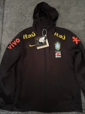  Brasilien vindjacka  - Svart träningsjacka från Nike med Brasilien landslagslogga på bröstet. Jackan har huva, dragkedja och tryck i gult, rött och grönt med sponsorloggor som Itaú och Vivo. Perfekt för fotbollsträning eller som supporterplagg.