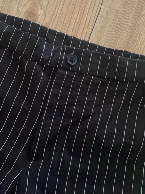 Svarta kostymbyxor med vita ränder - Säljer ett par svarta kostymbyxor med smala vita kritstrecksränder. Byxorna har raka ben, hög midja och stängs med knapp och dragkedja. Klassisk och stilren look som passar perfekt till en dressad outfit.