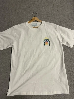 Vit t-shirt från Casablanca - Vit t-shirt från Casablanca med ett färgglatt tryck på bröstet som föreställer ett fönster med utsikt mot en byggnad och texten 'Avenida Casablanca' i gult. Klassisk passform och rund halsringning.