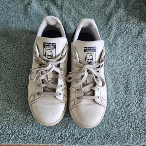 Adidas Stan Smith vita sneakers - Klassiska vita Adidas Stan Smith sneakers med blå detalj på hälen och perforerade ränder på sidorna. Skorna har lågt skaft, vita snören och är tillverkade i läder. Perfekta för en clean och tidlös stil.