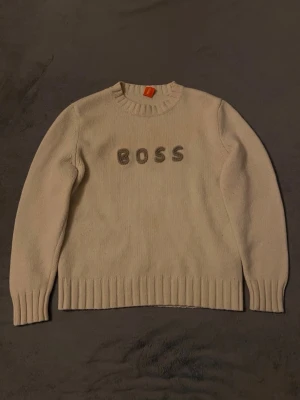 Beige stickad tröja från BOSS - Säljer en beige stickad tröja från BOSS med broderad logotyp på bröstet. Tröjan har rund hals, långa ärmar och ribbade muddar vid ärmslut och nederkant. Perfekt för dig som gillar stilrena plagg med diskret branding.