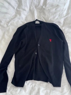 Svart cardigan från Ami Paris - Stilren svart cardigan från Ami Paris med röd broderad logga på bröstet. Tröjan har v-ringning, knäppning framtill och ribbade muddar. Perfekt att slänga över en t-shirt för en clean look.