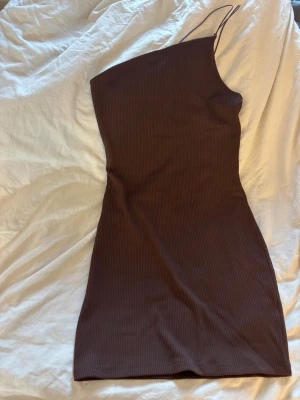 Brun one shoulder ribbad klänning - Säljer en brun, ribbad kortklänning med one shoulder-design och tunna axelband. Klänningen har en tight passform och är perfekt för dig som gillar en enkel men trendig stil. Materialet känns mjukt